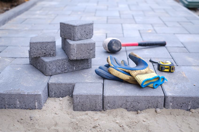 Gray paving stones being installed, with work gloves, a rubber mallet, and a tape measure placed on the ground.