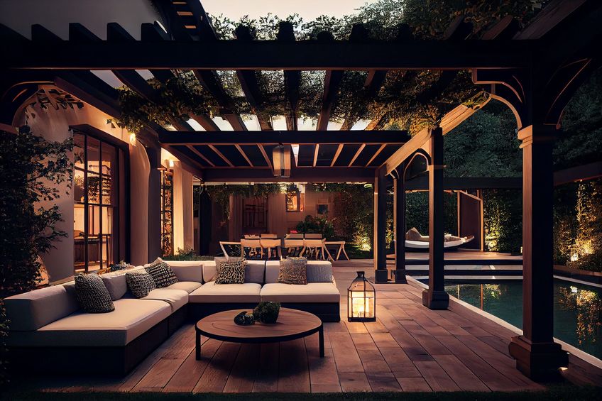 Modern outdoor patio with a pergola, sectional sofa, round table, lantern, dining area, and greenery, lit in the evening.