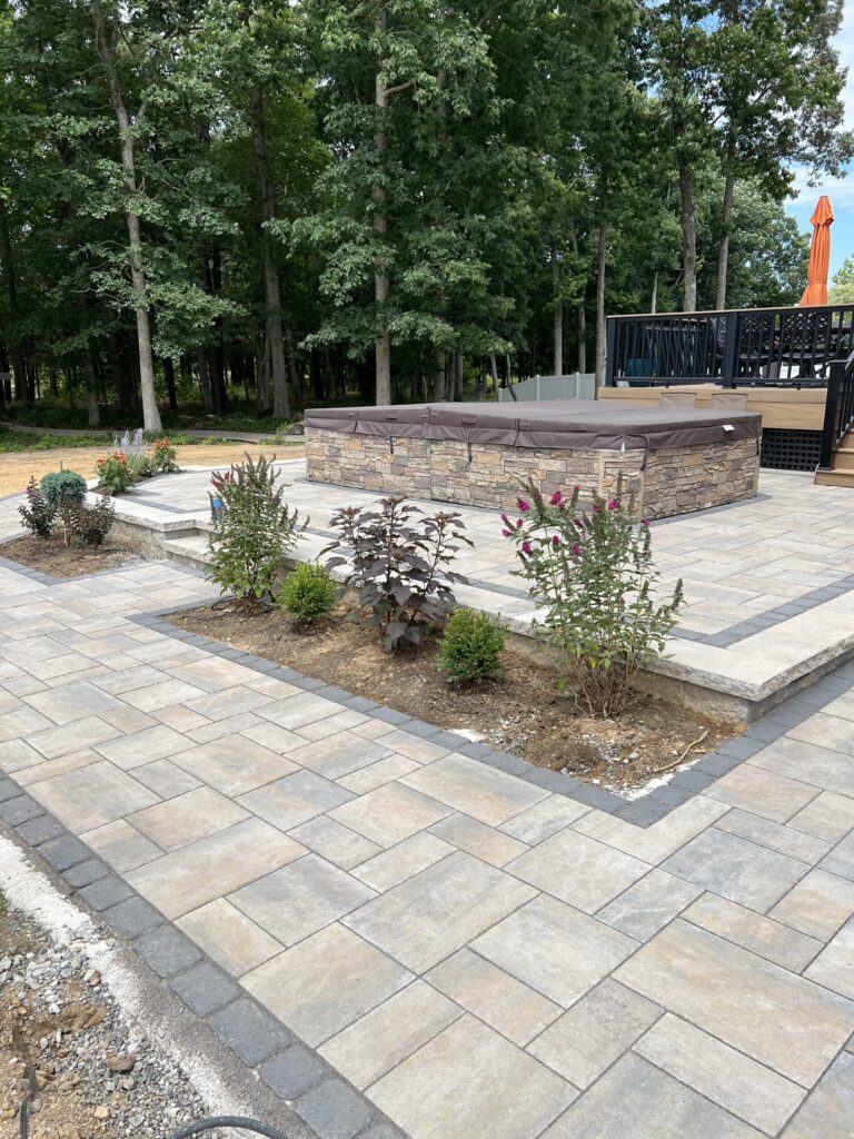 Stone patio with raised hot tub, garden beds with small plants, and surrounding trees in the background.
