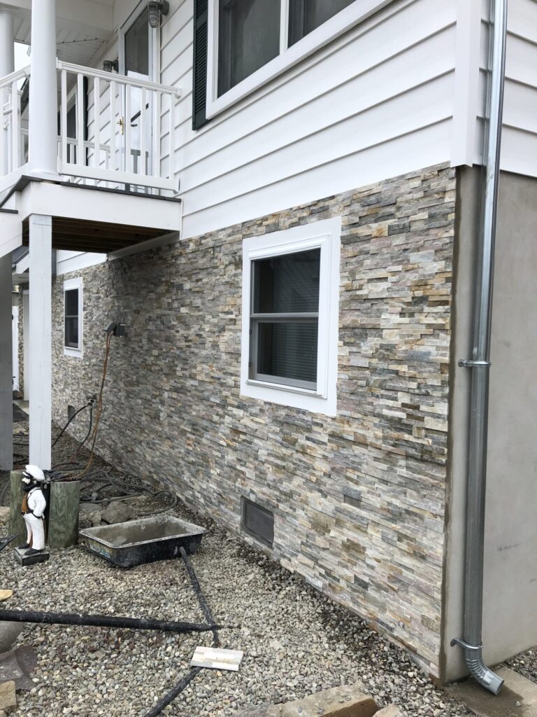 Stone veneer siding installed on the lower exterior wall of a house with white upper siding and a balcony above.