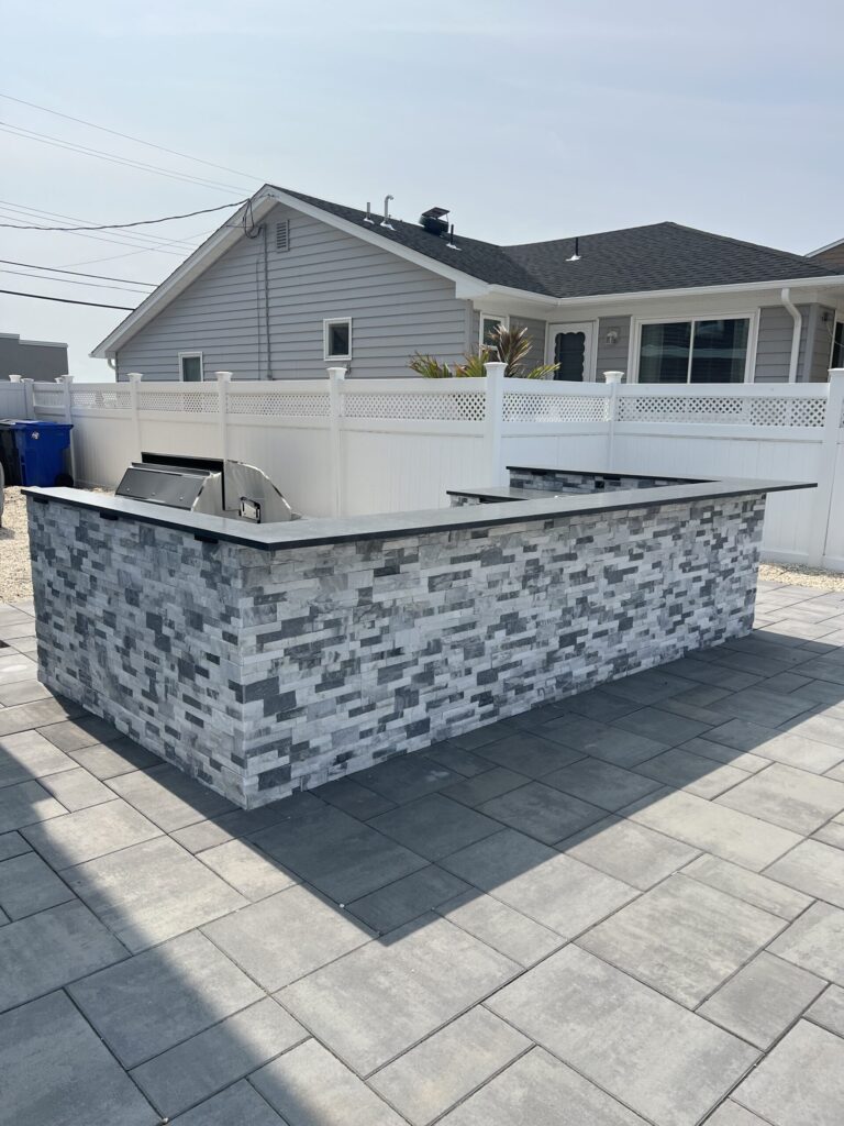 Outdoor kitchen island with gray stone veneer and built-in grill, set on a patio with concrete pavers.