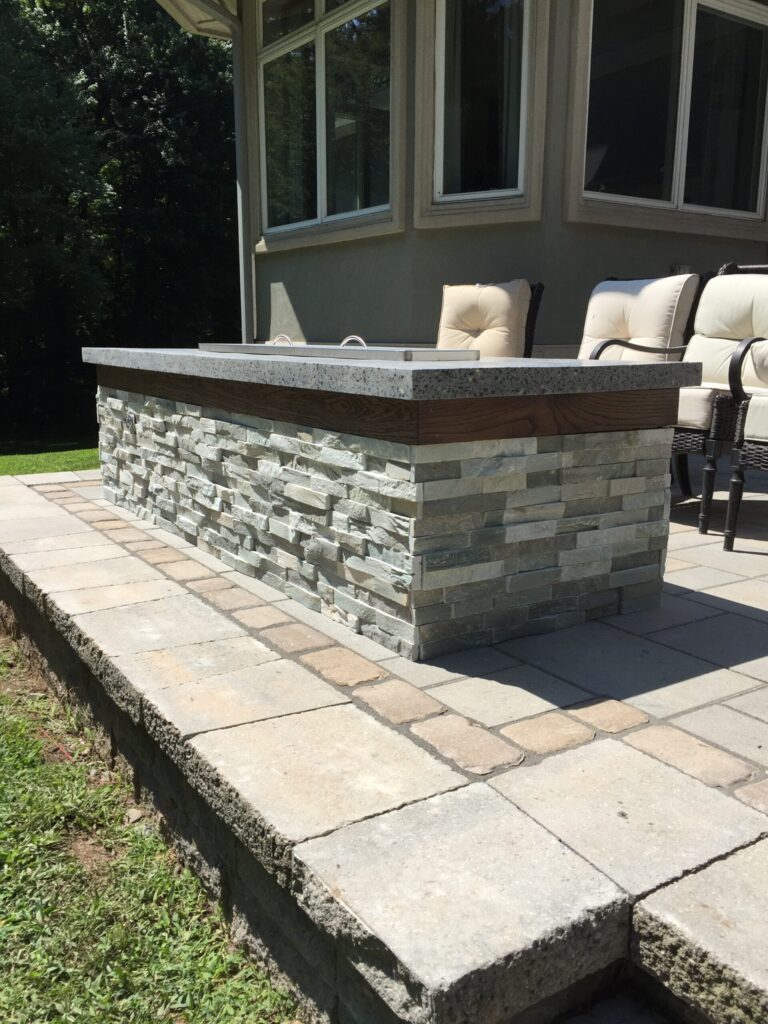 Rectangular stone outdoor firepit with a concrete top on a paved patio, next to cushioned patio chairs.