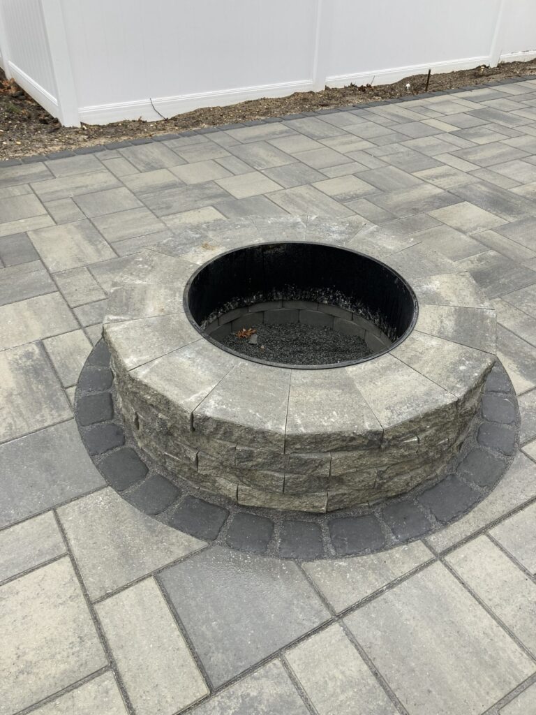 Round stone fire pit on a gray paver patio, bordered by a white fence and some bare ground.