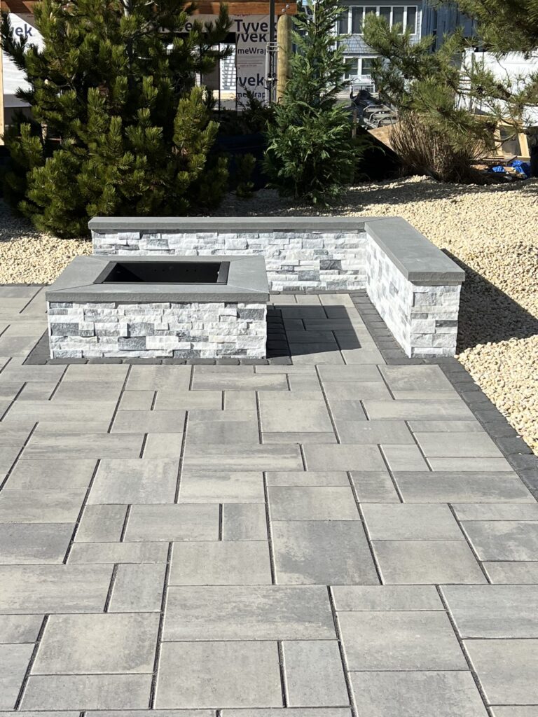 Gray stone patio with a rectangular fire pit and corner bench, surrounded by gravel and green shrubs.
