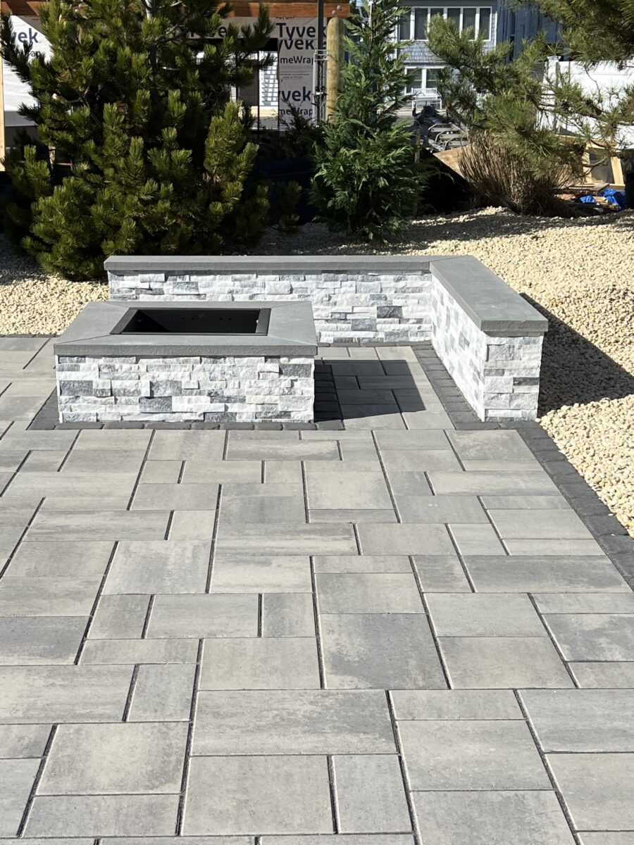 Gray stone patio with a rectangular fire pit and corner bench, surrounded by gravel and green shrubs.