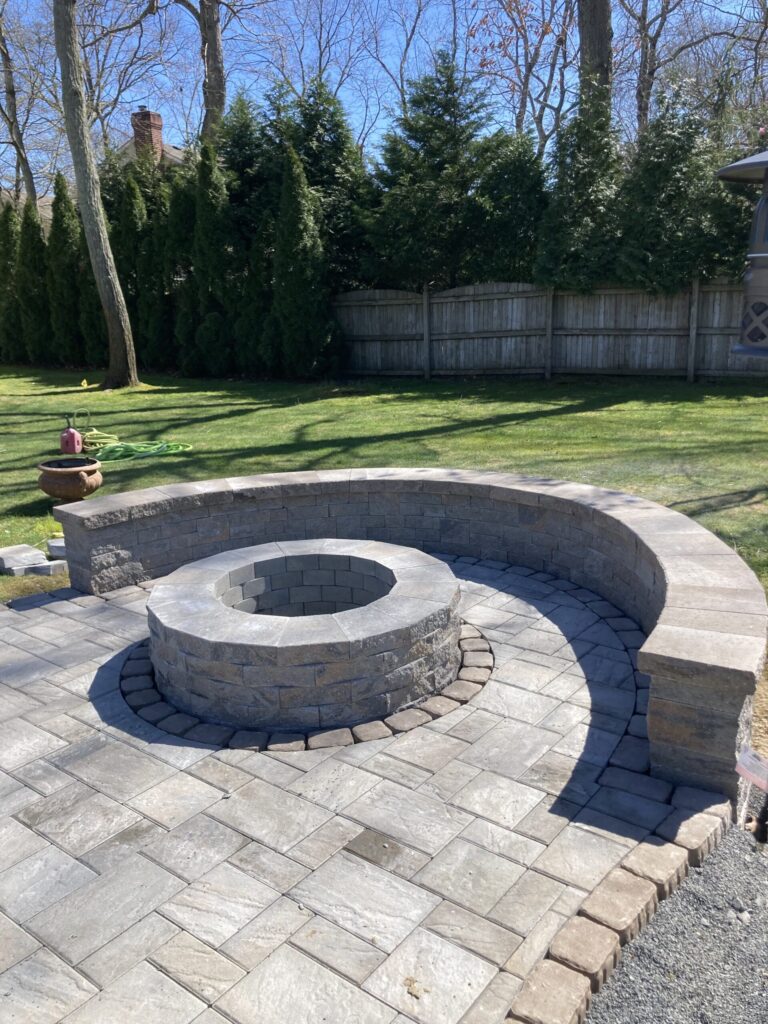 Circular stone fire pit with a curved stone bench on a paved patio in a fenced backyard with green grass and trees.