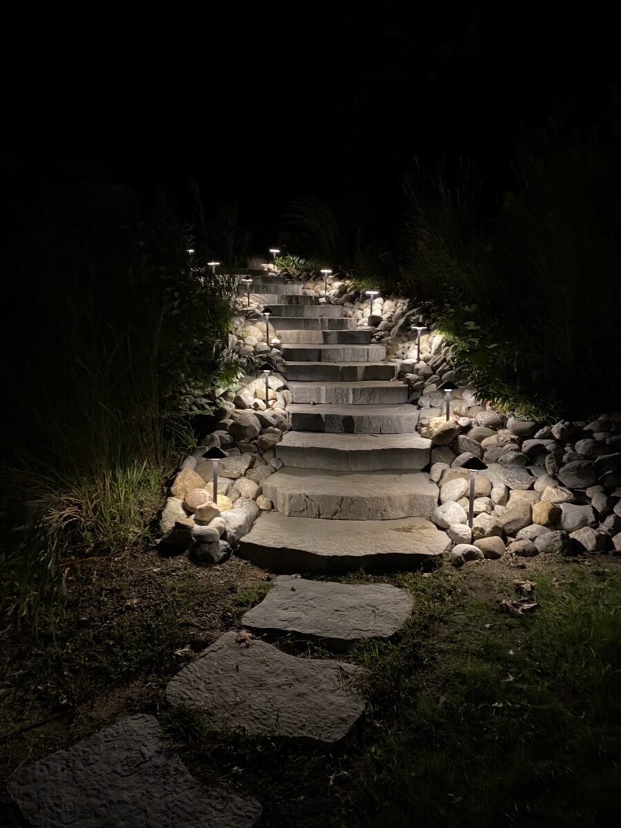 Stone steps with pathway lights ascend through rocks and plants at night, partially illuminated in the darkness.