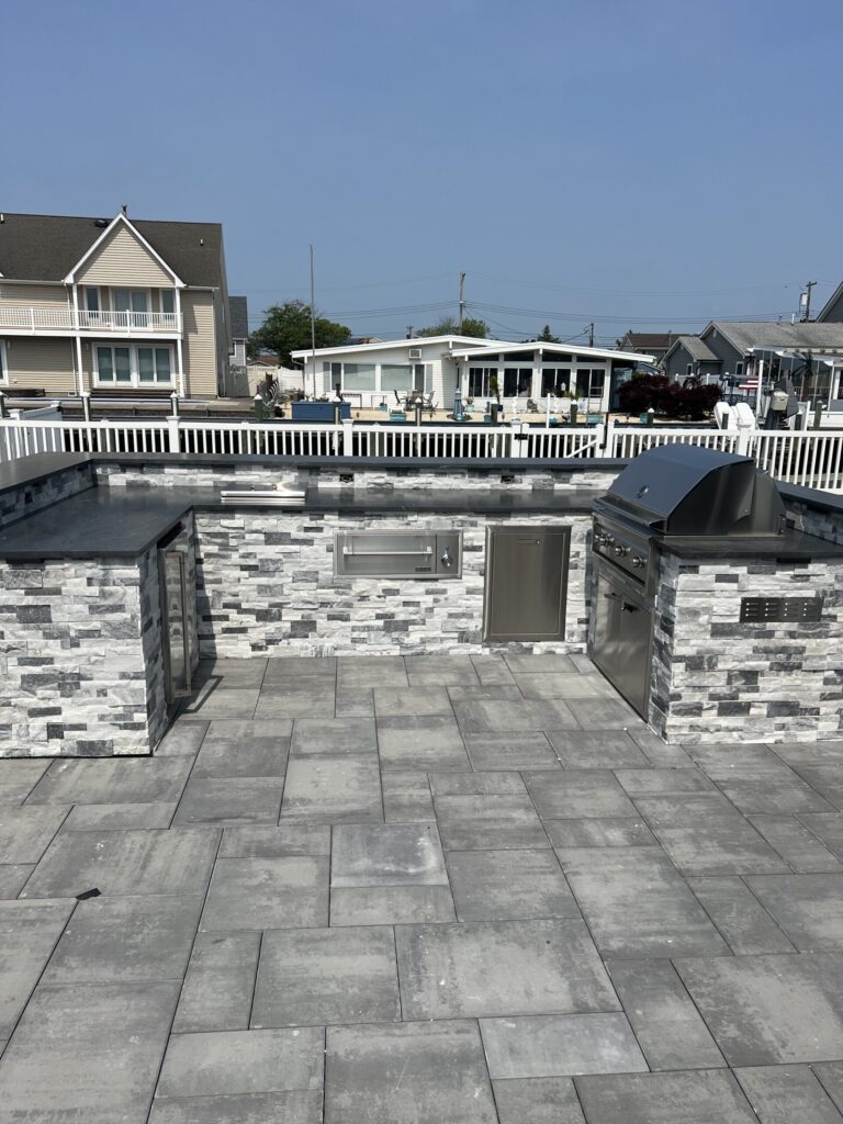 Outdoor kitchen with stone countertops, stainless steel grill, and appliances on a paved patio, surrounded by houses.