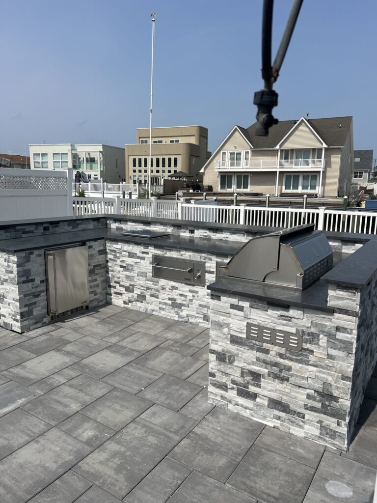 Outdoor kitchen with built-in grill, appliances, and stone finish on a paved patio, houses visible in background.