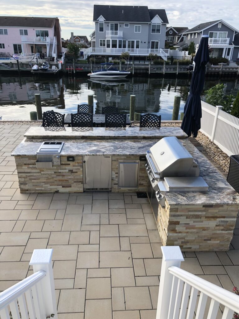 Outdoor kitchen with grill and bar seating overlooks a canal with houses, boats, and docks in the background.