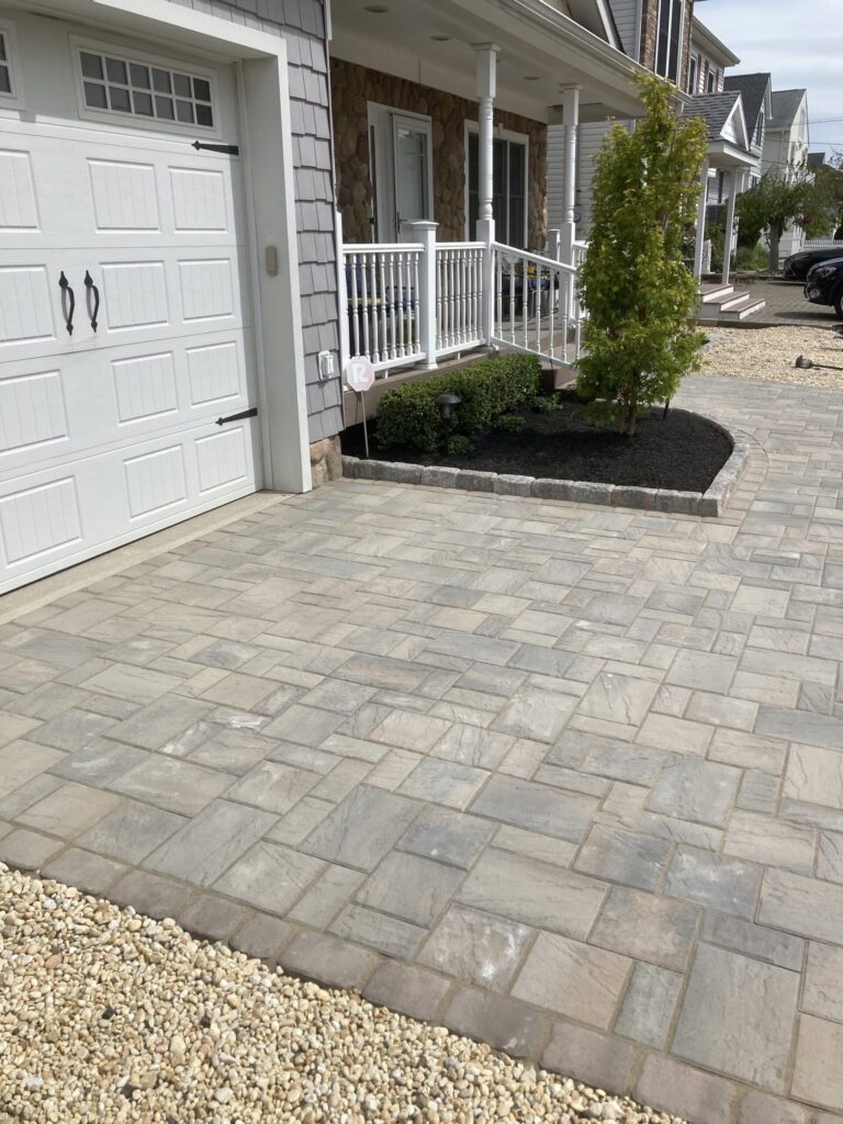 Driveway with patterned stone pavers next to a garage and porch with a small landscaped garden and shrubs.