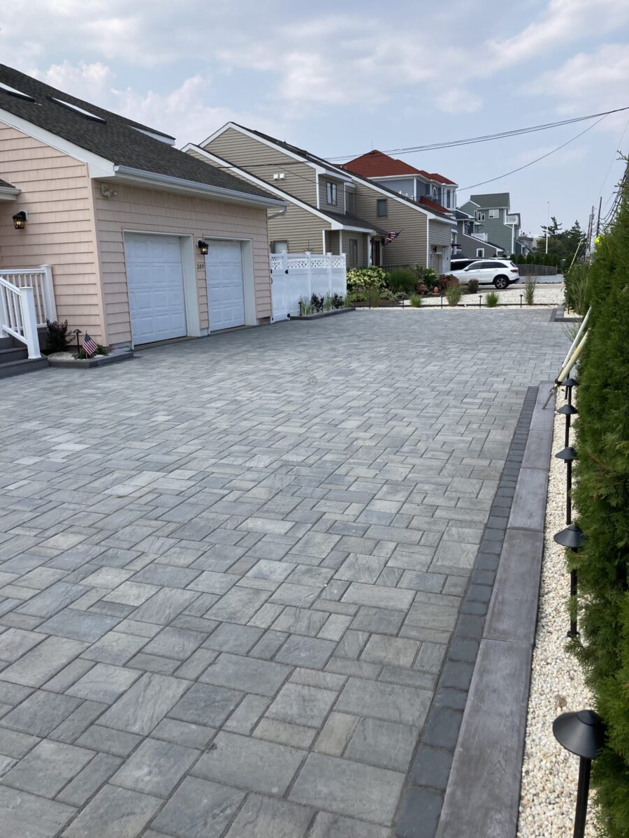 Brick-paved driveway leading to a double garage, with houses and greenery along the sides under a cloudy sky.