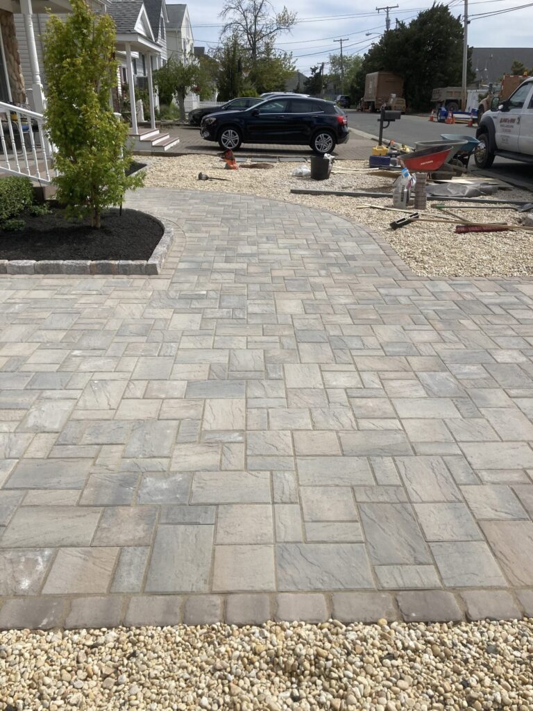 A paved stone walkway leads to a driveway with cars parked nearby and landscaping in progress.