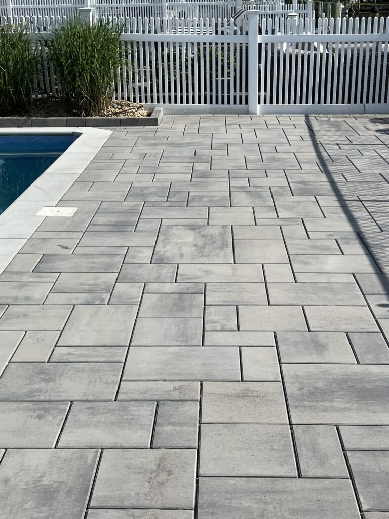 Gray rectangular pavers arranged in a geometric pattern beside a pool, with a white picket fence in the background.