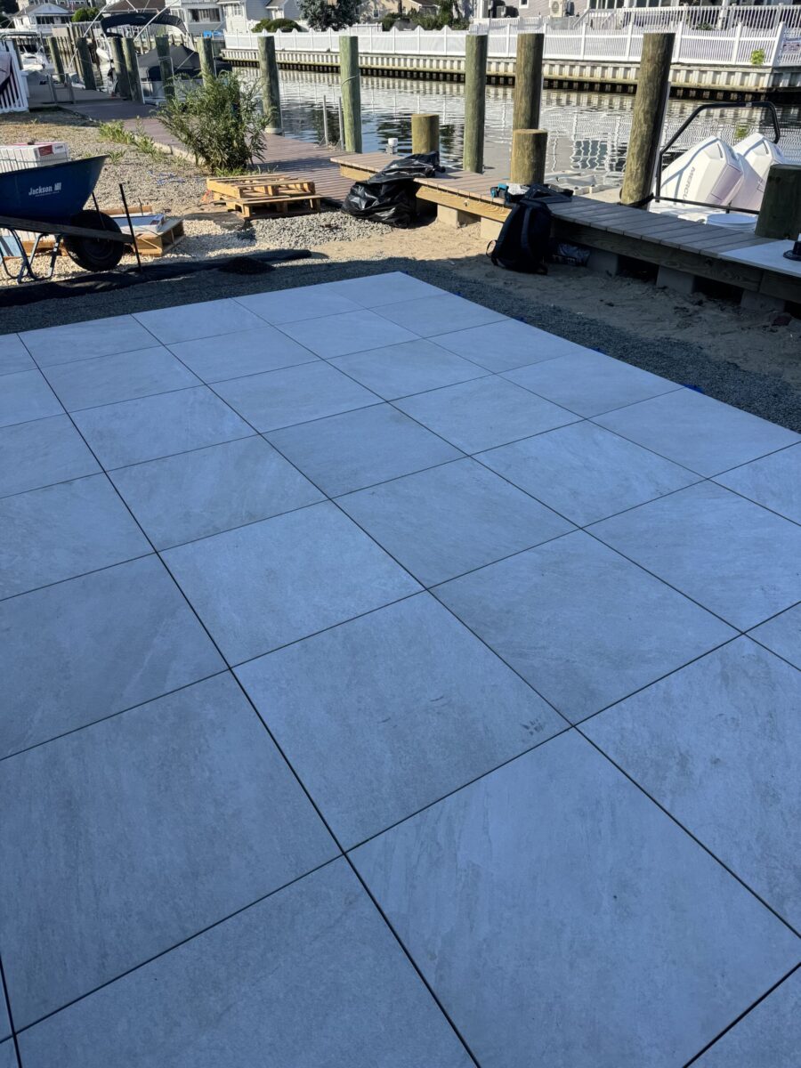 Square light gray tiles are newly installed outside near a wooden dock and a body of water.