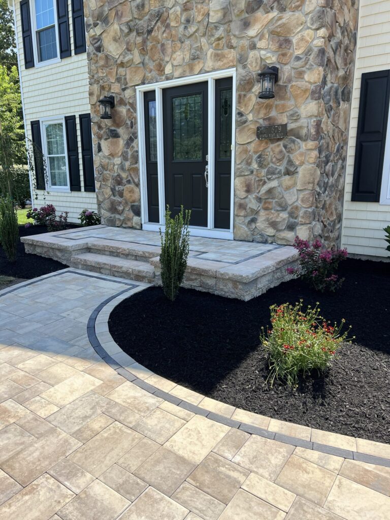 Stone house entrance with double doors, stone steps, lantern lights, paver walkway, and landscaped flower beds.