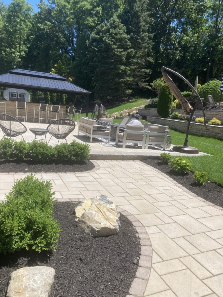 Patio with outdoor seating, fire pit, and umbrella, surrounded by landscaped garden and trees on a sunny day.