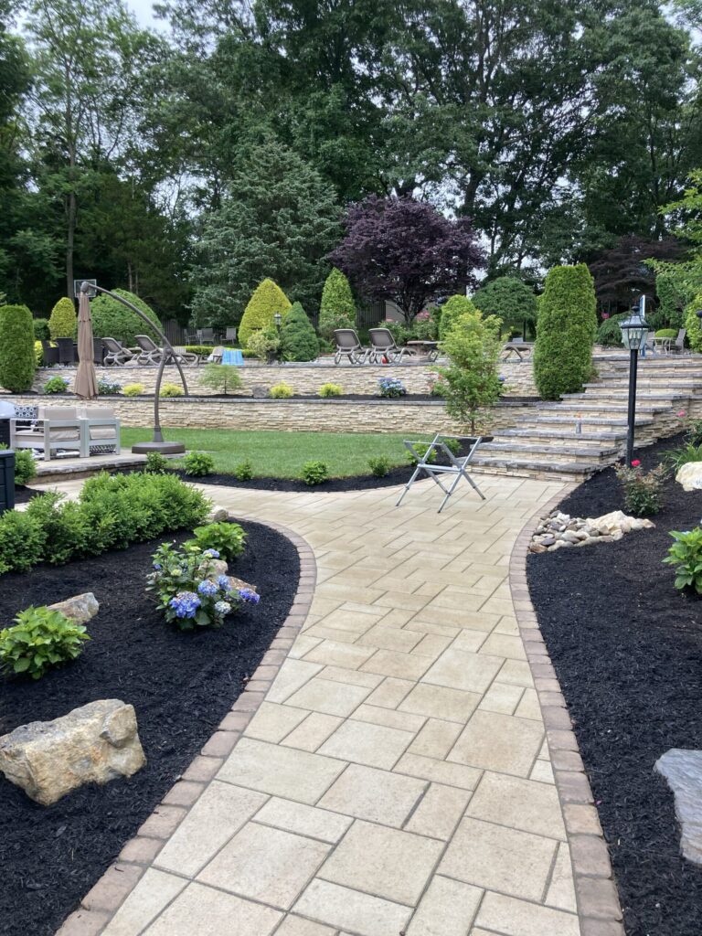 Paved walkway curves through landscaped garden with shrubs, flowers, and stone steps leading to patio area and pool.