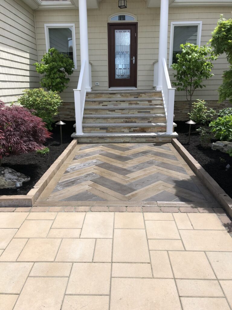 Stone walkway with a herringbone pattern leads to steps and a dark wooden front door, flanked by greenery.
