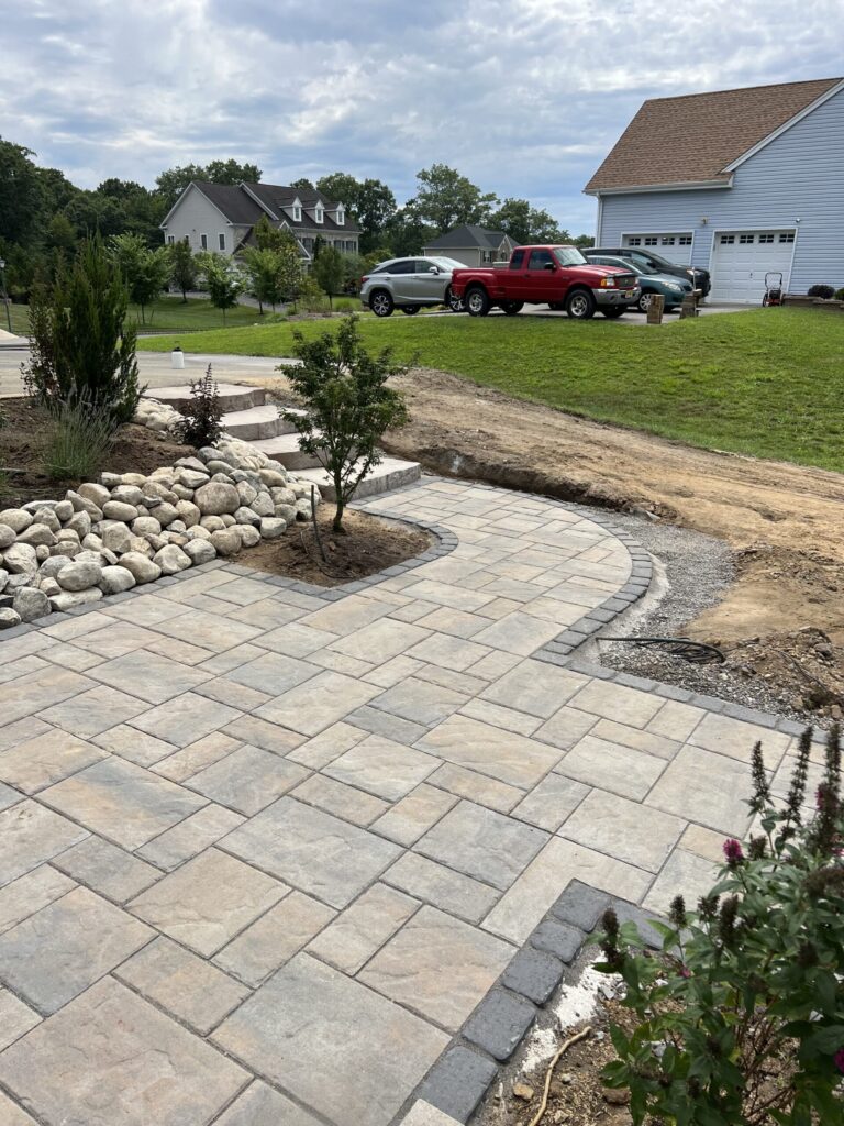Stone patio with a curved walkway, landscaping rocks, small plants, and parked vehicles in a suburban neighborhood.