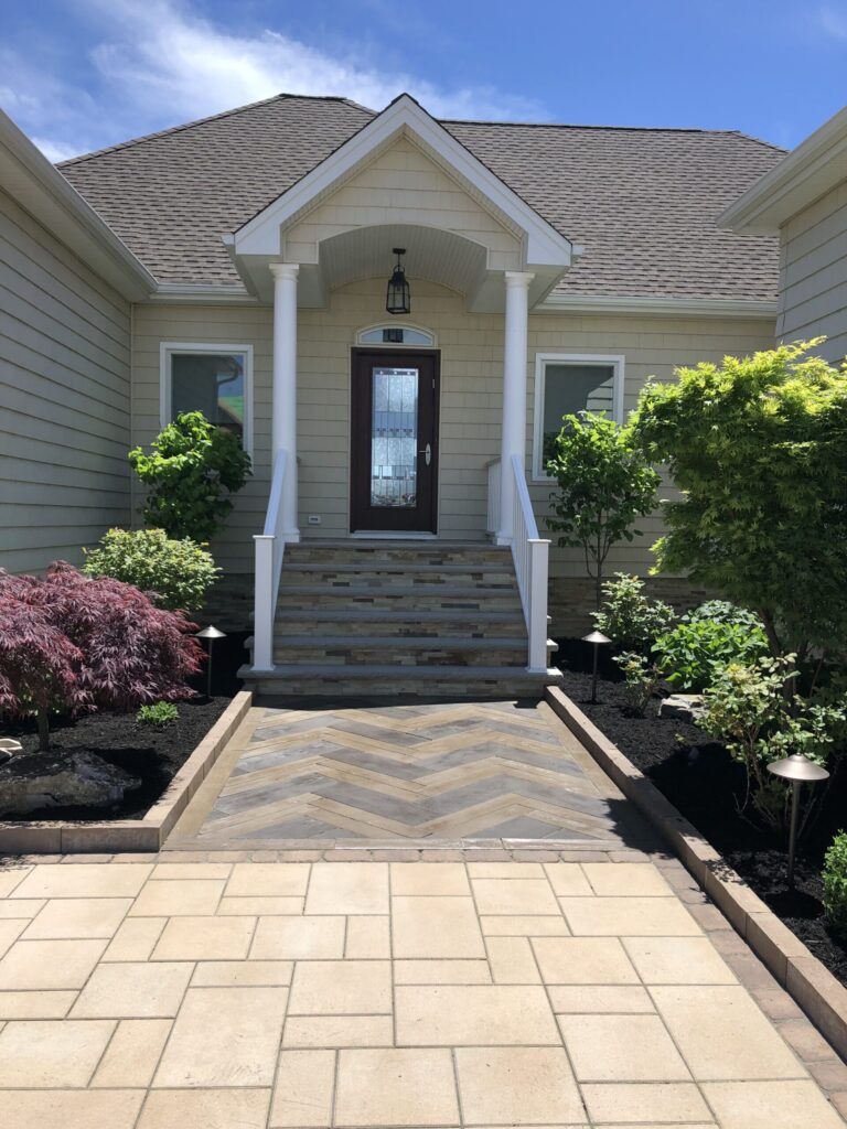 Front entrance of a house with a wooden door, white columns, steps, and landscaped plants on both sides of the walkway.