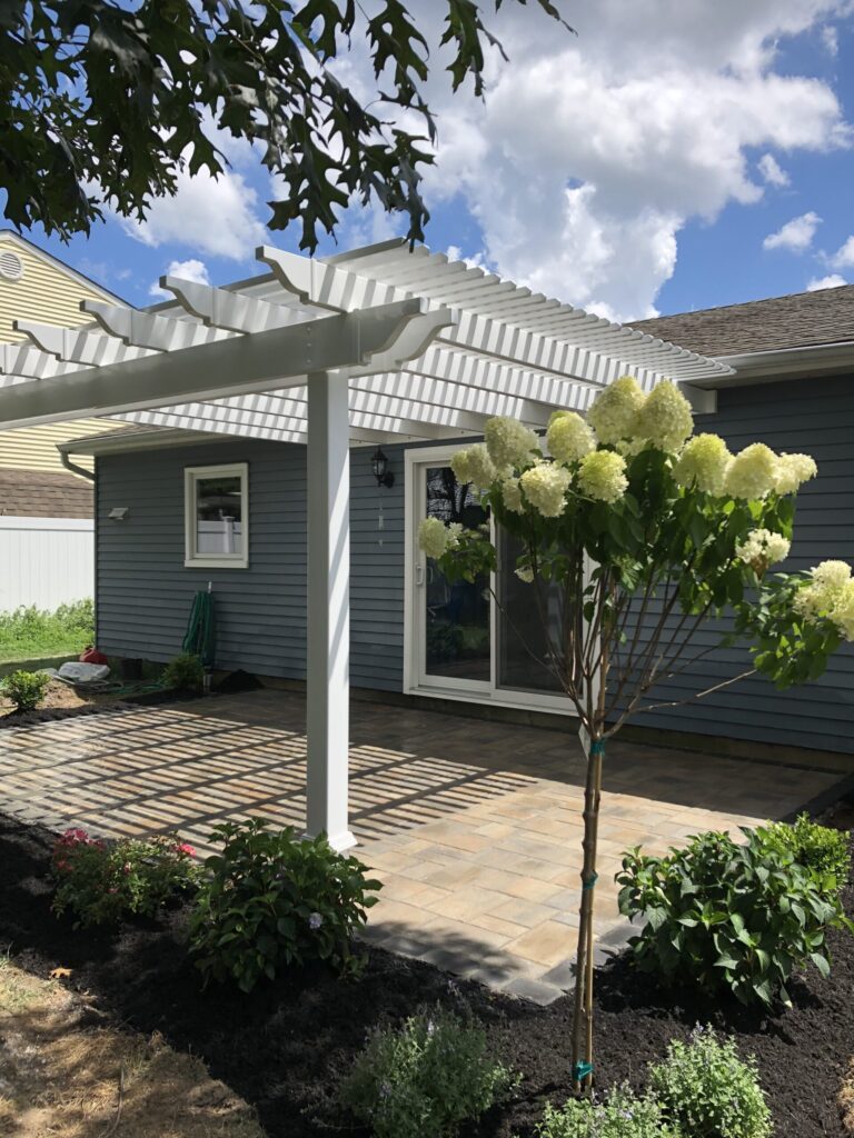White pergola over a patio with pavers, surrounded by shrubs and a blooming hydrangea tree next to a blue house.