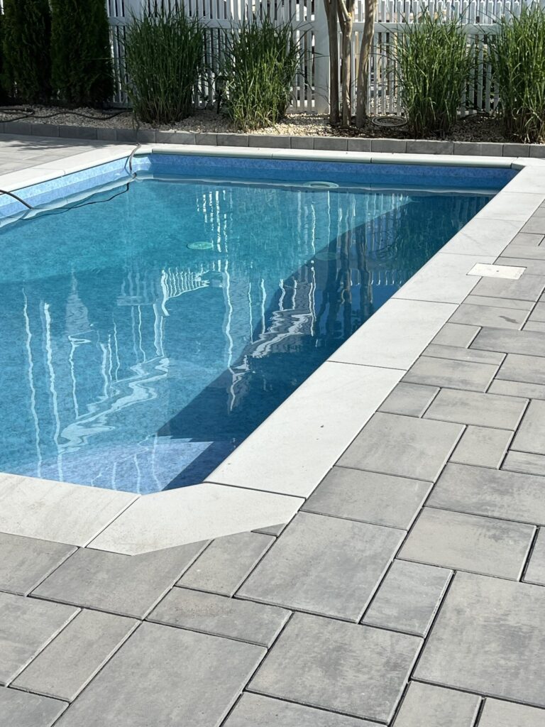 Rectangular outdoor swimming pool with clear blue water, surrounded by gray stone pavers and greenery in the background.