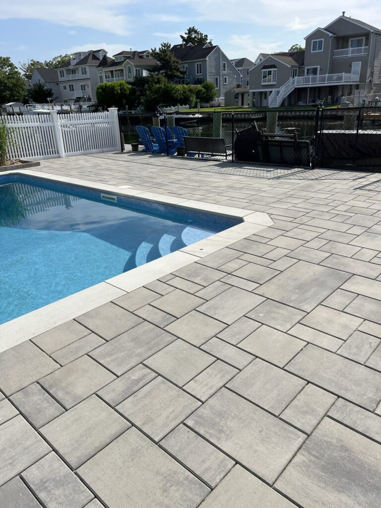 Rectangular outdoor pool with gray stone patio, white fence, and houses in the background under a blue sky.
