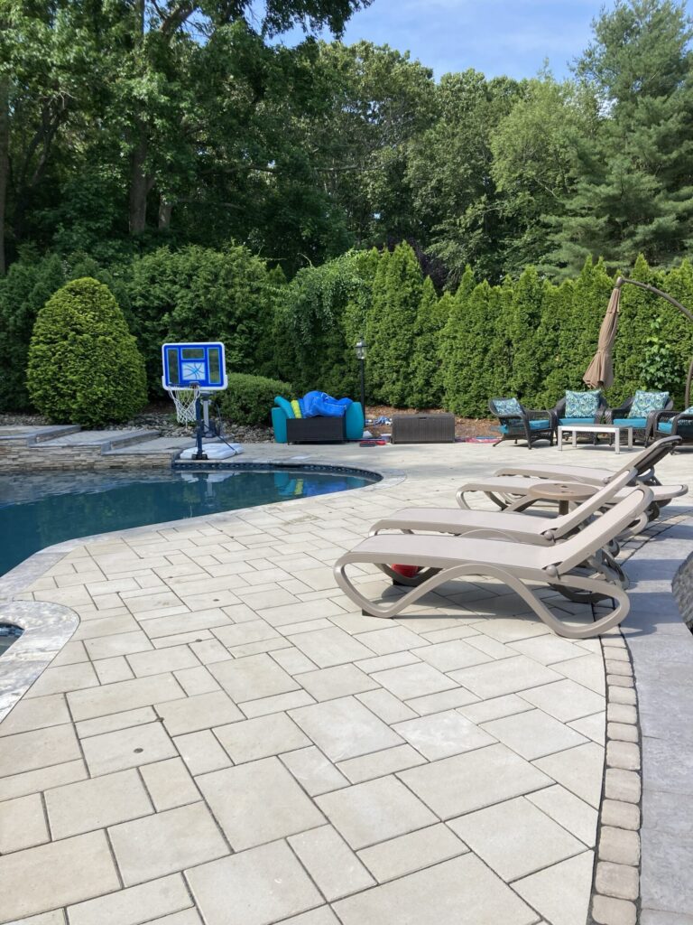 A backyard pool area with lounge chairs, a basketball hoop, patio furniture, and trees in the background.