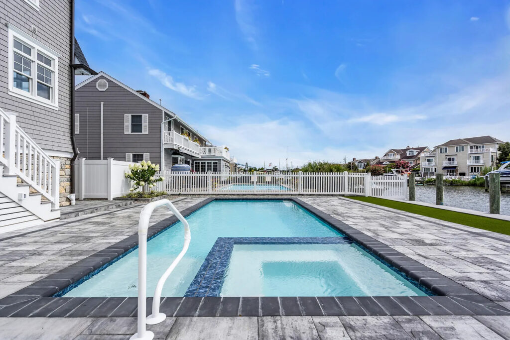 Rectangular backyard pool with built-in hot tub, surrounded by stone patio, near waterfront homes and docks.